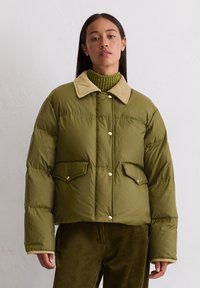 Olivgrüne Pufferjacke mit cremefarbenem Kragen, zwei Fronttaschen und Druckknöpfen. Getragen über einem grünen gerippten Rollkragenpullover und Cordhosen.