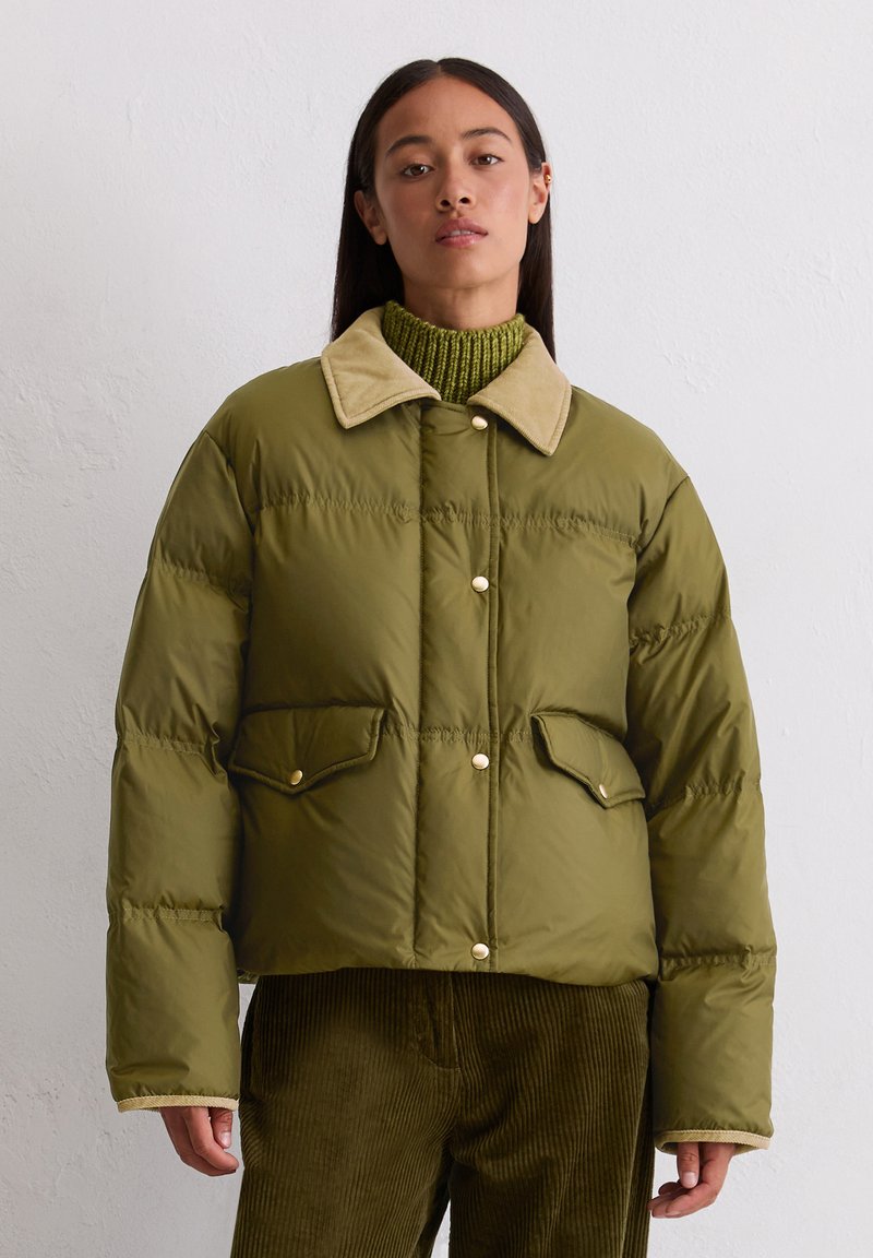 Olivgrüne Pufferjacke mit cremefarbenem Kragen, zwei Fronttaschen und Druckknöpfen. Getragen über einem grünen gerippten Rollkragenpullover und Cordhosen.
