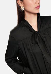 Schwarze Bluse mit einem Halsband aus Schleife, langen Ärmeln und plissiertem Stoff. Runde goldene Ohrringe setzen einen Kontrast mit einer spiegelnden Oberfläche.
