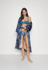 Roupa de noite longa em satin azul escuro com padrões florais, apresentando um forro interno rosa claro e um laço na cintura. Modelo usa um sutiã azul e roupa íntima de renda.
