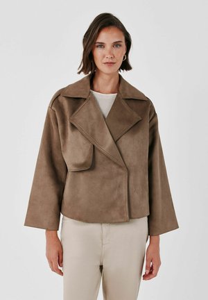 Femme aux cheveux bruns courts portant une grande veste en daim marron et un pantalon crème, debout devant un fond clair uni.
