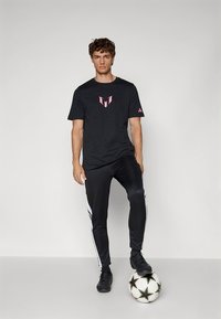 Tricou sportiv negru cu logo roz, asortat cu pantaloni negri cu dungi laterale albe, purtat cu pantofi negri. Minge de fotbal la picioare.