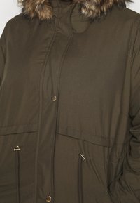 Parka verde olivo con capucha forrada de piel sintética, dos bolsillos frontales, cinturilla ajustable con cordón y cierres de botón de metal. Textura de tejido suave.