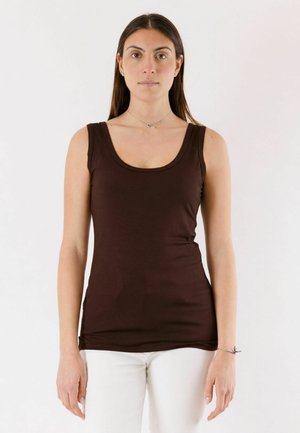 Donna in piedi, fronte rivolta in avanti, che indossa un top marrone scuro senza maniche e pantaloni bianchi contro uno sfondo bianco uniforme.