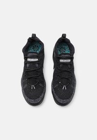 Svarta atletiska skor med en texturerad stickad ovansidan, reflektiva detaljer, runda snören och en synlig dämpad sula. Inuti har de ett färgglatt mönster.