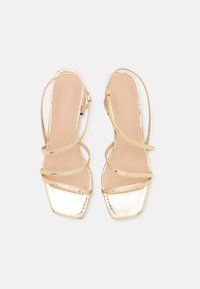 Sandales dorées à lanières avec un bout carré, présentant une finition métallique texturée et de fines lanières. La semelle intérieure est de couleur beige clair.