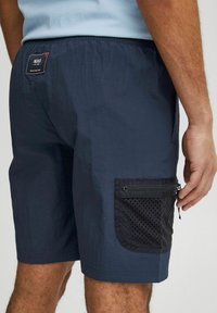 Shorts bleumarin dintr-un material ușor, cu un buzunar lateral din plasă neagră pentru ventilație și un mic eticheta cu logo pe spate.