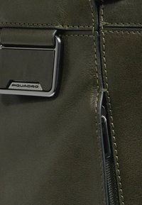 Borsa in pelle verde oliva con accessori neri, caratterizzata da una chiusura lampo prominente e una targhetta con logo rettangolare. Dettaglio della superficie testurizzata visibile.