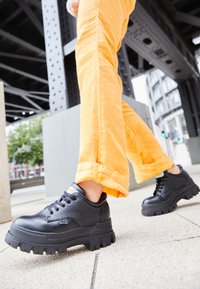 Sneakers platform in pelle nera con suole in gomma spessa, punta tonda e design classico con lacci, abbinate a pantaloni arancioni con risvolto.