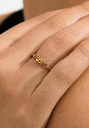 Bague en or présentant un design en écriture cursive "maman", posée sur une main avec une texture de peau visible, mettant en valeur une finition métallique lisse et une forme délicate.
