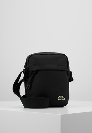 Borsa a tracolla nera con tracolla regolabile e piccolo logo verde a forma di coccodrillo nell'angolo anteriore, posizionata su una superficie bianca su sfondo grigio.