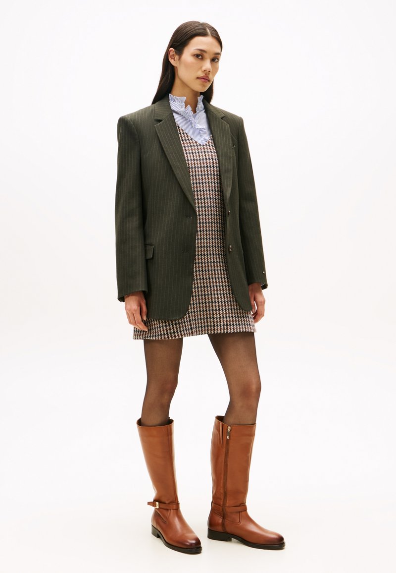 Brune lærstøvler til knæene, grønn stripet blazer, kjole med hundetannmønster og blå stripete skjorte med krage. Glatte teksturer.