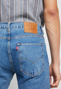Blå denimjeans med en läderlapp som har varumärkesinformation. Distinkt bakficksyddning och en röd etikett på fickan.