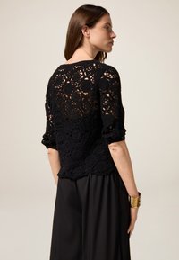 Svart virkade topp med geometriska mönster, ovala former och en texturerad yta. Har trekvartslånga ärmar och en lös passform.