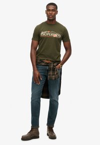 Groen katoenen t-shirt met grafische print, gecombineerd met blauwe denim jeans en bruine veterschoenen. Een geruite blouse om de taille gebonden voegt textuur toe.