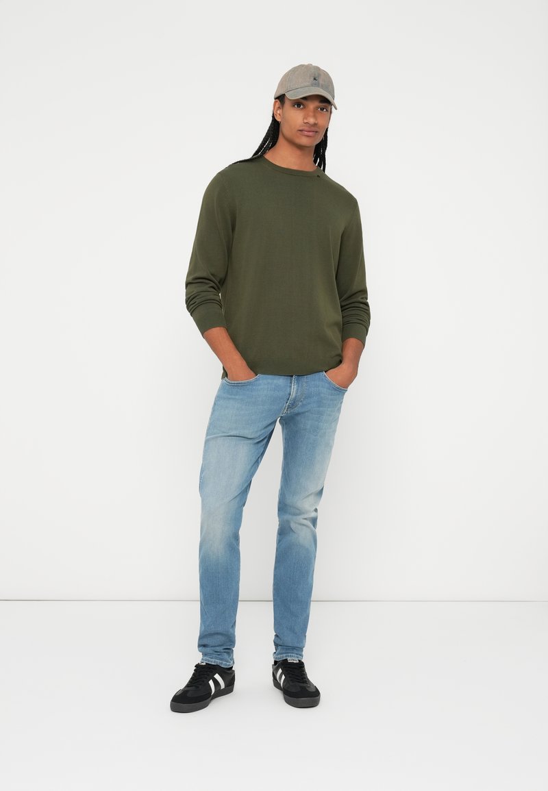 Olivgrüner Pullover mit langen Ärmeln, hellblaue Jeans, schwarze Sneakers mit weißen Streifen und eine beige Mütze. Einfaches, figurnahes Design.