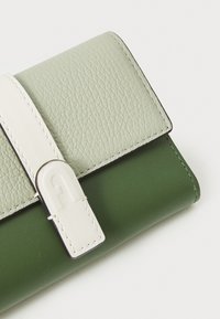 Portofel din piele verde și gri deschis, cu o suprafață texturată, o curea albă și un sistem de închidere din metal argintiu. Design minimalist cu culori contrastante.