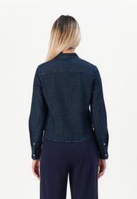 Denim jacket in dark blue with a fitted design, pointed collar, long sleeves, and buttoned cuffs. Smooth texture and curved hem.
