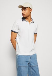 Tricou alb cu guler și manșete contrastante în nuanța bleumarin, asortat cu blugi largi de culoare bleu deschis. Modelul poartă o șapcă bej.