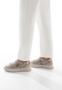 Persona con pantaloni bianchi e sneakers beige con lati testurizzati e foderati in pelliccia, in piedi su una superficie bianca.