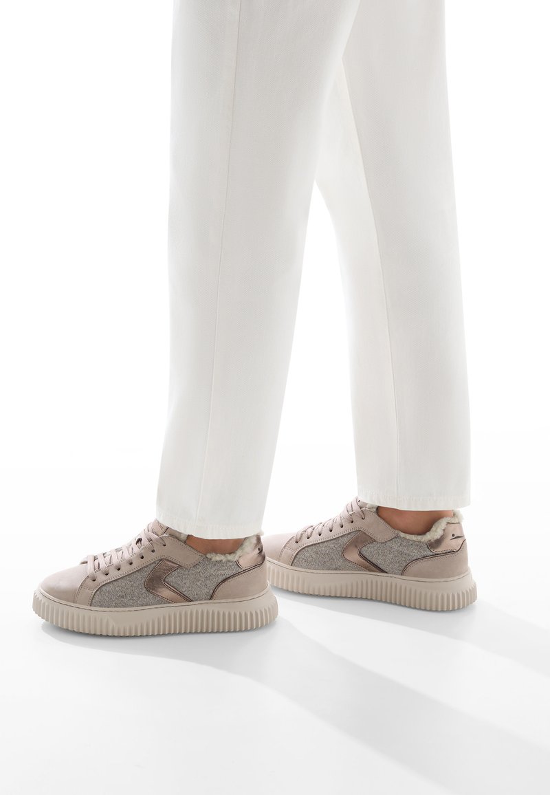 Persona con pantaloni bianchi e sneakers beige con lati testurizzati e foderati in pelliccia, in piedi su una superficie bianca.