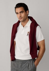 Biely polo tričko s logom na hrudi, doplnené vínovým sveterom prehodeným cez plecia a šedými károvanými nohavicami.