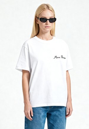 Blond vrouw met zwarte zonnebril, wit T-shirt met korte mouwen en de tekst "Mira Paris", en een blauwe spijkerbroek tegen een effen lichte achtergrond.