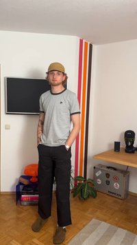 Siva majica s crnim obrisom, nosi se s crnim cargo hlačama i smešnim cipelama. Stojeći u sobi s zidovima ukrašenim šarenim prugama.
