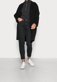 Černý dlouhý kabát přes úzký černý rolák, doplněný o černé džíny s vysokým pasem a bílé tenisky. Hladké materiály a pohodlný střih.