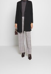 Sorte lang frakke over en grå turtleneck, parret med vide ternede bukser i grå og beige, samt spidse brune ankelstøvler. Holder en lille taske.