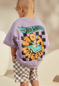 Übergroßes Fliederfarbenes T-Shirt mit schwarzem und orangefarbenem "Hot Wheels"-Grafik, das ein türkisfarbenes Auto zeigt, kombiniert mit karierten grauen Shorts.