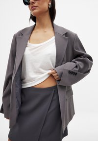 Grå oversize blazer med en enkelknäppt design, vit lös skjorta under och en matchande grå kjol. Svarta knappdetaljer är synliga.