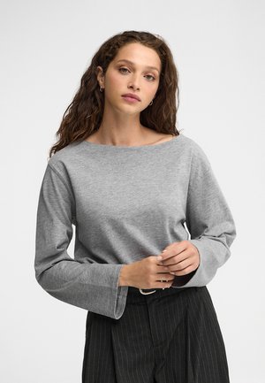 Mujer con cabello castaño ondulado que lleva pequeños pendientes de aro, una camiseta gris de manga larga y pantalones negros de rayas finas con cinturón sobre un fondo blanco.