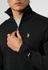 Približen posnetek osebe, ki zadrguje temno sivo jopo z majhnim vezenim emblemom in tricolor zastavo na manšeti rokava.