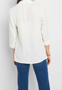 Vit blus med trekvartsärmar, slät tyg, rundad fåll och knäppta manschetter, i kombination med blå denimbyxor.