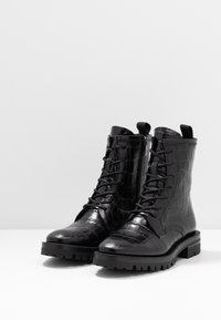 Un par de botas negras de cuero con estampado de cocodrilo, con cordones y cremalleras laterales, y suelas resistentes sobre una superficie blanca.