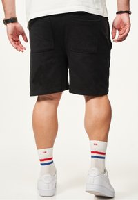 Schwarze Baumwollshorts mit zwei Gesäßtaschen, kombiniert mit weißen Turnschuhen und mehrfarbigen Socken in Rot, Weiß und Blau.