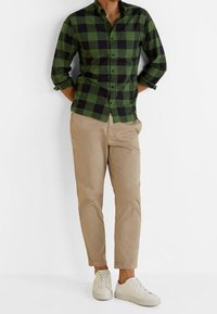 Uomo che indossa una camicia a scacchi verde e nera, pantaloni beige e sneakers bianche, in piedi con le mani dietro la schiena.