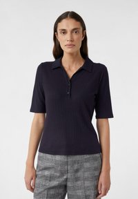Granatowa koszulka polo z prążkowanego materiału, z krótkimi rękawami, zapięciem na trzy guziki i płaskim kołnierzykiem. W połączeniu z szarymi spodniami w teksturowany wzór w kratę.