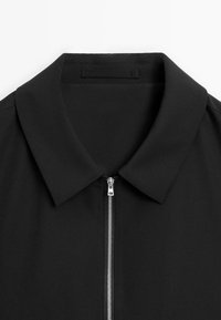 Black collared shirt with a front zipper. Smooth fabric texture and minimalist design. Features a sharp, folded collar and subtle stitching.