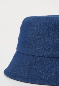 Sombrero tipo bucket de denim en azul oscuro, con un logo bordado en la parte frontal, textura suave y un ala ancha que se inclina hacia abajo.
