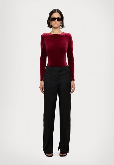Burgunderfarbener Samt-Bodysuit mit langen Ärmeln, kombiniert mit schwarzen, hoch taillierten Hosen mit seitlichen Schlitzen und einer taillierten Passform. Schwarze Sonnenbrille getragen.
