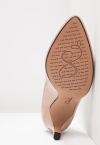 Talons hauts en cuir verni nude avec un bout pointu, semelle texturée arborant un logo et une finition lisse et brillante. Accent noir au bout du talon.