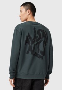 Tmavě zelená mikina vyrobená z bavlny, s velkými černými nápisy na zadní straně, které zní "ALLSAINTS LONDON." Obsahuje žebrované manžety a lem.