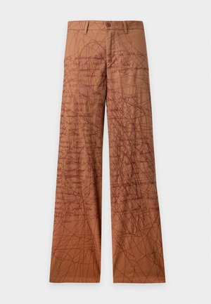 Homme portant une casquette rouge, une chemise crème et un pantalon marron avec des motifs abstraits en lignes rouges, debout les mains dans les poches sur un fond uni.