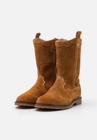 Ein Paar braune Wildlederstiefel in Knöchelhöhe mit runden Spitzen, dekorativen Nähten und Zuglaschen auf einem weißen Hintergrund.