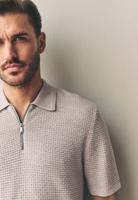 Polo à manches courtes beige clair avec un motif texturé et un col zippé, présenté sur un fond neutre.