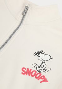 Haut à fermeture éclair blanc présentant une illustration de Snoopy en cartoon et le texte "SNOOPY" en lettres roses grasses. Tissu lisse avec un détail de fermeture éclair élégant.