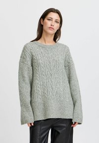 Hellgrauer, gestrickter Pullover mit einem Kabeldesign, weiten Ärmeln und einem runden Ausschnitt. Weiche Textur und lässige Passform, kombiniert mit dunklen Hosen.