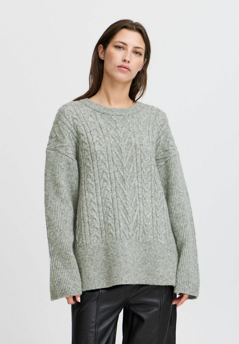 Hellgrauer, gestrickter Pullover mit einem Kabeldesign, weiten Ärmeln und einem runden Ausschnitt. Weiche Textur und lässige Passform, kombiniert mit dunklen Hosen.
